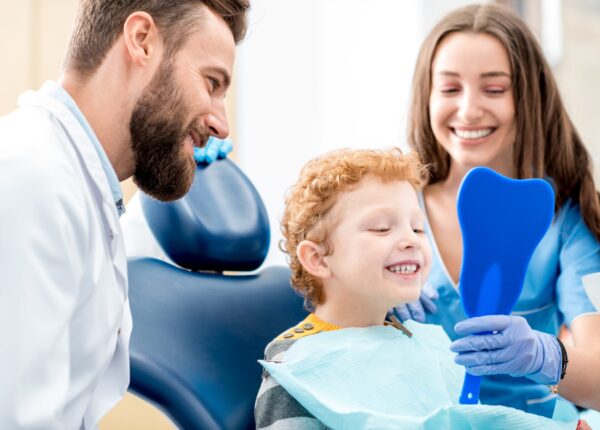 Young boy looking at the mirror with toothy smile sitting on the chair with dentist and assistant at the dental office